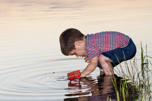 Un niño de 3 años juega en la orilla del río con juguetes brillantes, cava arena. Juegos para niños cerca del estanque al aire libre en el verano. — Foto de Stock