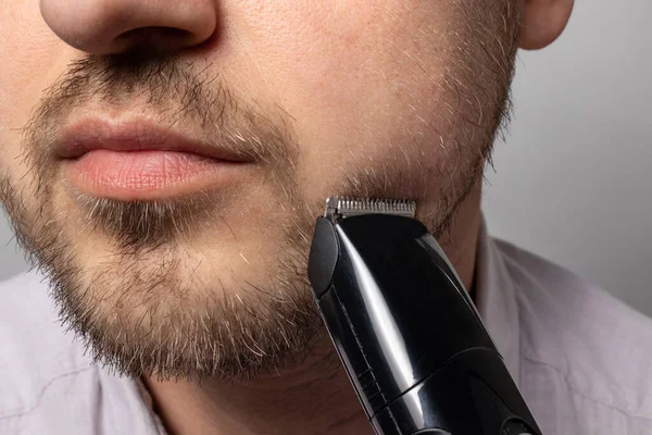 A man shaves his beard with a trimmer razor. Modeling beard, masculine style, facial hair care, morning routines in the bathroom.