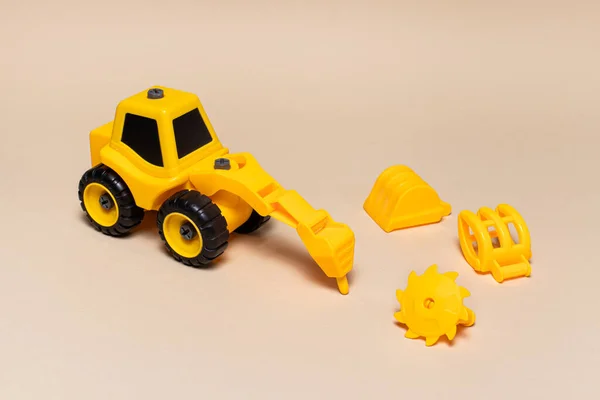 Giocattolo di plastica da bambini su uno sfondo beige - un trattore giallo con diversi ugelli trapano, sega su legno, una pala caricatrice di legno, un escavatore a benna. Attrezzature da costruzione per bambini. — Foto Stock
