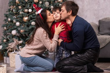 Anne ve baba Noel ağacının yanında küçük bir çocuğu öpüyor. Ailenle yeni yılın kutlu olsun. Ebeveynler ve çocuklar