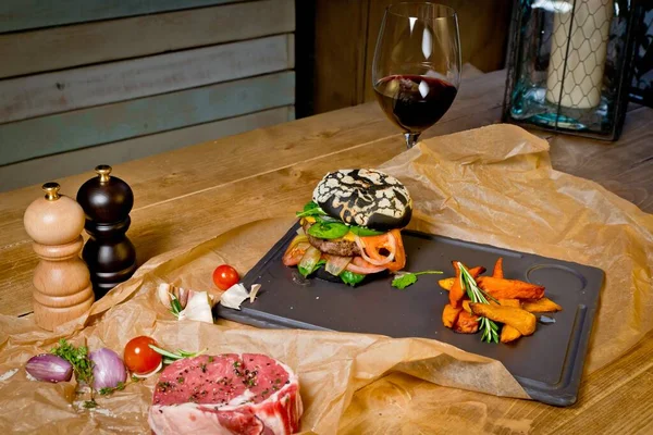 Black beef Burger with fresh garlic and potatoes and a glass of red wine. On a black plate. On a rustic wooden table — Stock Photo, Image