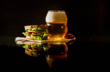 Bir bardak bira ve peçetedeki peynirli, sosisli ve salatalı sandviç siyah arka planda yansıması olan cam bir masanın üzerinde.