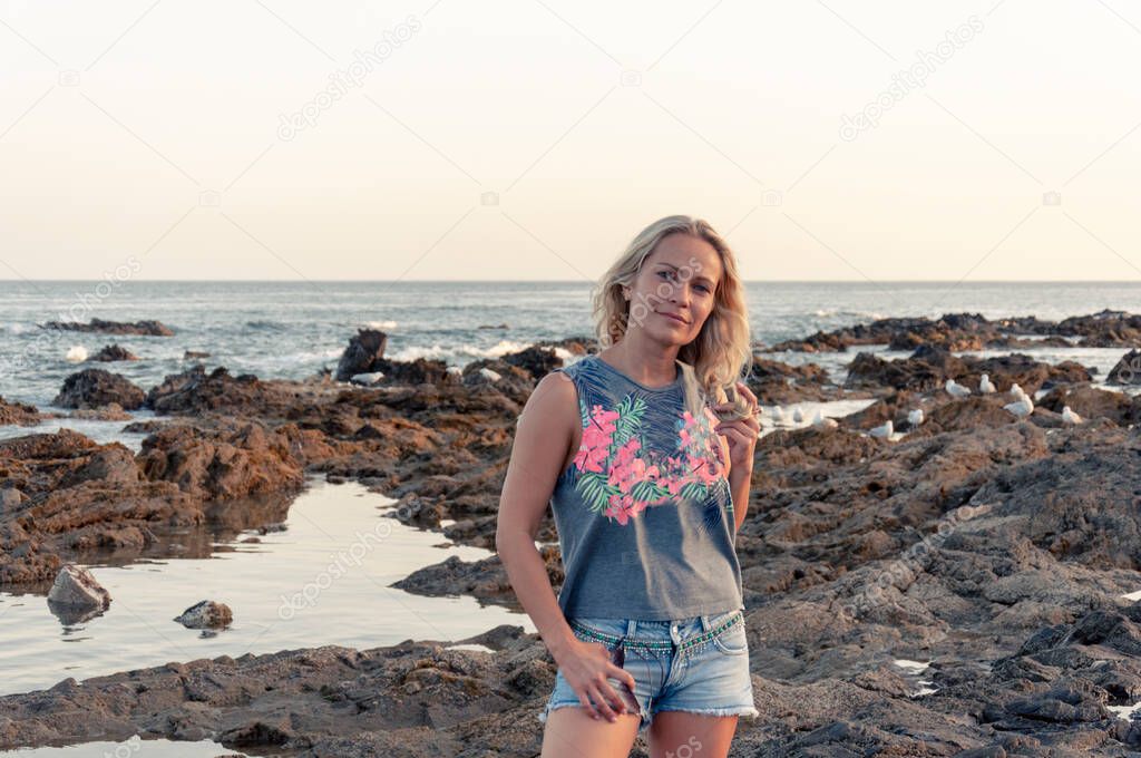 Girl dreamily looks into the lens with her head slightly tilted to the side against the background of the sea and the rocky shore