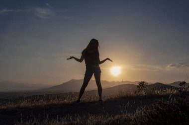 Siyah tişörtlü, kot şortlu ve şapkalı bir kızın silueti elleri tepenin üstünde, gün batımının ve dağların arkasında duruyor.