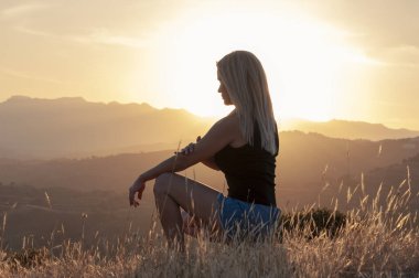 Tişörtlü, şortlu ve spor ayakkabılı sarışın bir kızın, batan güneşin ve dağ zirvelerinin arka planında otların arasında çömelmiş oturuşunun yan görüntüsü..