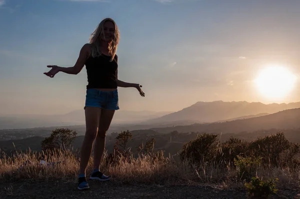 A girl in a black T-shirt, jeans shorts and hats, with her hands spread apart, stands on top of a hill against the backdrop of sunset and mountains.