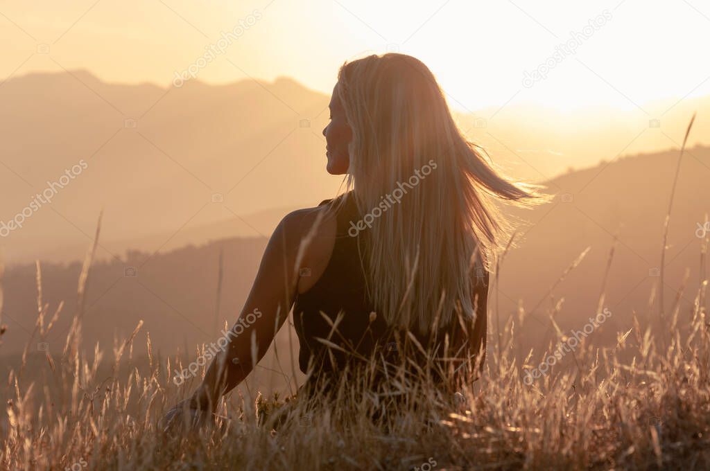 A girl sits in the grass on a hilltop with her eyes closed and meditates against the backdrop of mountain peaks and the sunset, her blond hair flying in the wind.