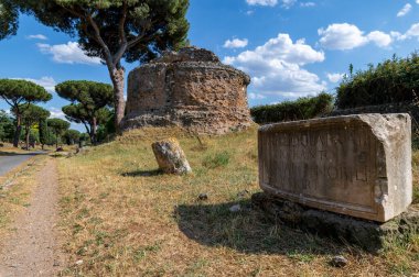 Güneşli bir günde Via Appia 'da dairesel tuğla mezar ve epigraf manzarası. Yol, Roma İmparatorluğu 'nun en önemli yoluydu. Diğer mezarların kalıntıları, zengin bitki örtüsü, deniz çam ağaçları, selvi ağaçları..