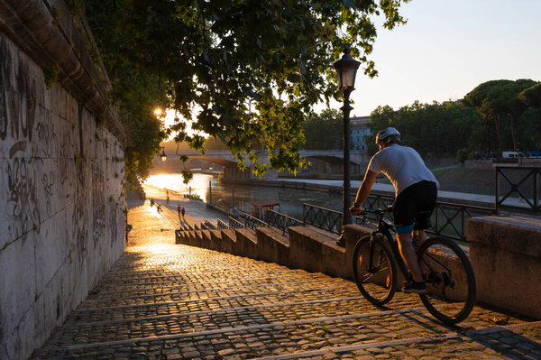 Walk along the Tevere, at sunset with the sun on a summer day near the Tiber island. A cyclist walks the staircase flooded by the rays of the sun that is setting in the Tevere. Rome, Italy.