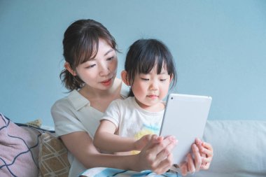 Anne ve kızı kanepede oturuyor ve tablet bir cihaz kullanıyorlar.