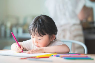 Asian girl drawing with colorful pens at the dining table at home clipart