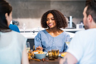 Kadın ve erkeklerin yemek masasını sardığı bir ev partisi sahnesi.