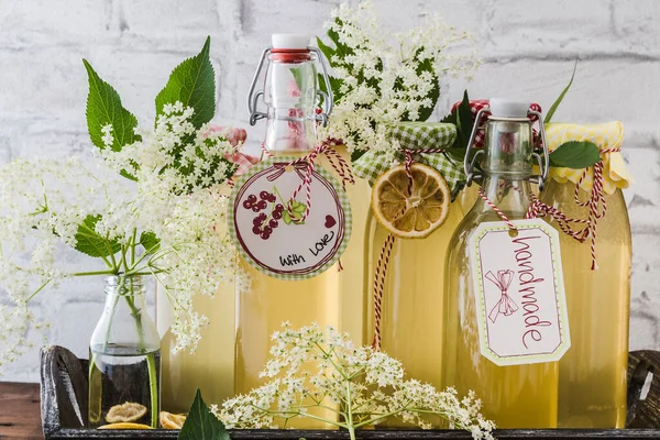 Different bottles with homemade elderflower syrup in front of a white wall, decorated with elderflowers and dried lemon slices