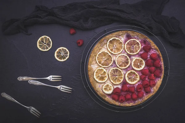 Cheese cake with raspberries, dried lemon slices, forks and black cloth on dark background, top view