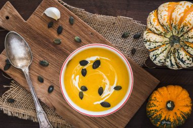 Krema ve balkabağı tohumlu balkabağı çorbası koyu kahverengi ahşap zemin üzerinde ahşap bir tahta üzerinde, küçük balkabakları, kaşık ve tohumlarla süslenmiş, üst görünüm.