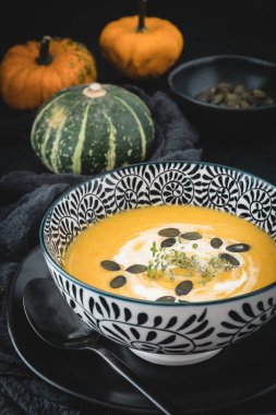 Siyah ve beyaz kâse, balkabağı çorbası kremalı ve siyah zemin üzerinde tohumlar, küçük balkabaklarıyla süslenmiş. Dikey depo fotoğrafı. 