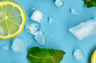 Mavi arka planda kesilmiş turunçgil meyveli kompozisyon. Limon dilimleri, nane yaprakları ve buz küpleriyle yaratıcı bir yaz kompozisyonu. Limonata içeceği konsepti. Üst görünüm.