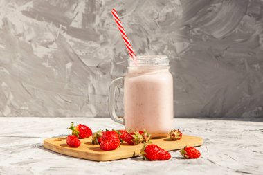 Cam bir kavanozda kırmızı çizgili bir tüple çilekli smoothie. Tahtada çilek var ve yanında da çilekli smoothie. Sağlıklı gıda kavramı. Hafif beton arka planda