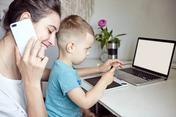 A woman with a child works at a computer and speaks on the phone. Concept of work from home and home family education. Mom and son are is working on a graphics tablet and on a laptop at home.