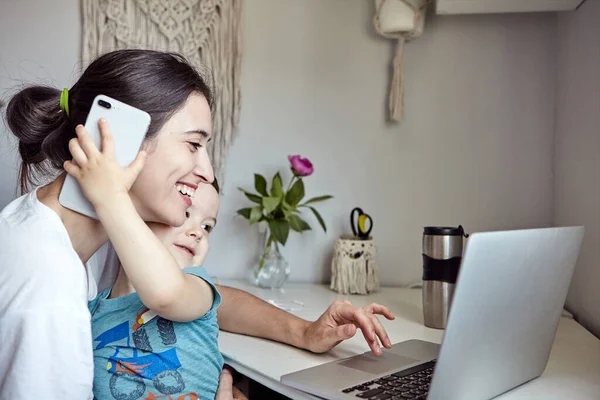 A woman with a child works at a computer and speaks on the phone. Concept of work from home and home family education. Mom and son are is working on a graphics tablet and on a laptop at home.
