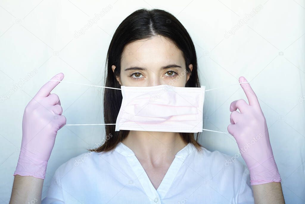 A woman in pink gloves puts on a pink protective mask. The girl stands on a light background in white clothes. The concept of suppression of coronavirus