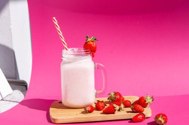 Cam bir kavanozda kırmızı çizgili bir tüple çilekli smoothie. Tahtada çilek var ve yanında da çilekli smoothie. Sağlıklı gıda kavramı. Parlak pembe bir arka planda