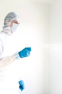 Koruyucu giysili eldiven ve maske takmış elinde sprey tutan bir adam. Antibakteriyel tedavi kavramı. Beyaz bir arkaplanda izole edilmiş