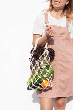Hafif yaz kıyafetleri içinde genç bir kadın elinde sebzelerle örgü alışveriş torbası, poşetsiz yeşillikler tutuyor. Sıfır atık, plastiksiz çevre dostu kavram. El yapımı makrome çanta. Sürdürülebilir yaşam tarzı.  