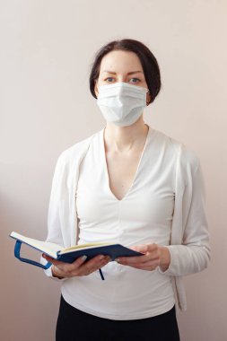 Koruyucu maskeli bir kadın beyaz bir ceketle arka planda duruyor ve elinde bir günlük tutuyor. Not alan bir tıp uzmanı.