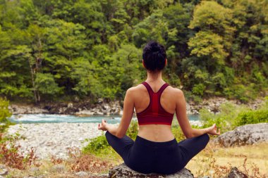 Doğada yoga dersleri. Tek başına spor yapma kavramı. Sosyal dışlama. Bir kadın kayalıklarda yoga yapıyor, bir dağ nehrinin yanında.