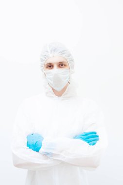 Koruyucu takım elbiseli ve yüz maskeli bir adam kollarını kavuşturmuş kameraya bakıyor. Beyaz bir arkaplanda izole edilmiş.