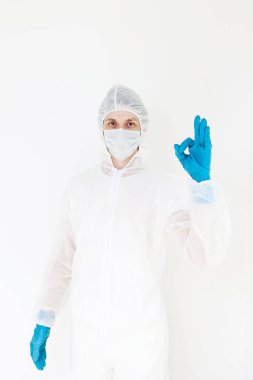 Koruyucu takım elbiseli ve yüz maskeli bir adam. Tamam jestini gösteriyor. Beyaz bir arkaplanda izole edilmiş