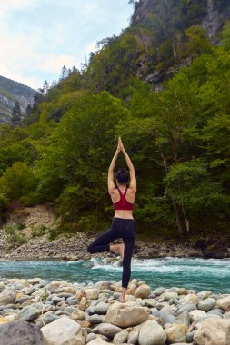 Doğada yoga dersleri. Tek başına spor yapma kavramı. Sosyal dışlama. Bir kadın kayalıklarda yoga yapıyor, bir dağ nehrinin yanında.