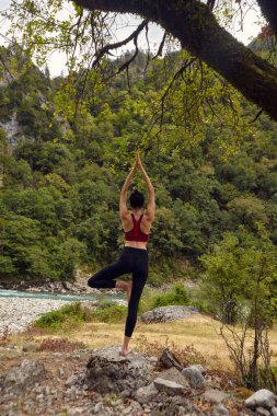 Doğada yoga dersleri. Tek başına spor yapma kavramı. Sosyal dışlama. Bir kadın kayalıklarda yoga yapıyor, bir dağ nehrinin yanında.
