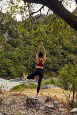 Doğada yoga dersleri. Tek başına spor yapma kavramı. Sosyal dışlama. Bir kadın kayalıklarda yoga yapıyor, bir dağ nehrinin yanında.
