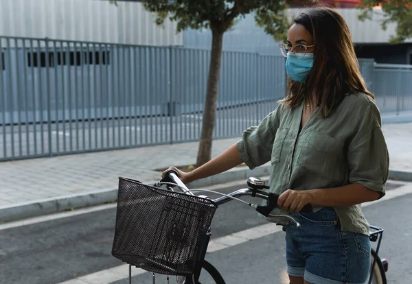 Young girl with glasses and a face mask is holding a bicycle in a urban atmosphere