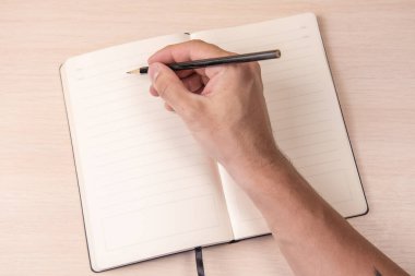 Ahşap arka plan görünümünde, elinde siyah kalemle açık bir defter, okul not defteri ve masada bir erkek eli, ofiste bir masaüstü..