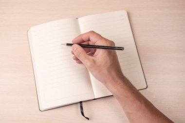 Ahşap arka plan görünümünde, elinde siyah kalemle açık bir defter, okul not defteri ve masada bir erkek eli, ofiste bir masaüstü..