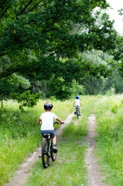Kasklı ve açık renk elbiseli iki çocuk güneşli bir günde parkta bisiklet sürüyor..
