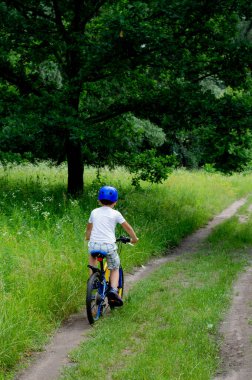 Kasklı ve açık renk elbiseli bir çocuk güneşli bir yaz gününde ormanda bisiklet sürüyor..