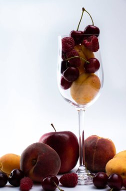 Şeffaf bir bardakta, kayısılar, ahududular, hafif arka planda kirazlar. Kırmızı elma ve şeftali.