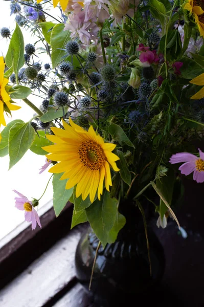 A bouquet of sunflower and other wild flowers in a dark vase on a windowsill on a sunny day.