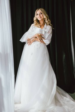 Düğünden önceki sabah, genç güzel sarışın gelin beyaz bir elbise deneyerek düğün törenine hazırlanıyor, romantik bir fotoğraf.