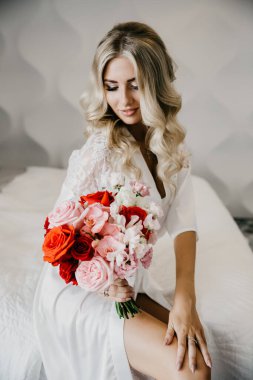 Düğün için. Beyaz stüdyo içinde, tıpkı evdeki gibi, kanepede oturan gül buketli gelin. Modaya uygun bir düğün fotoğrafı. Gelin olarak genç, çekici sarışın manken, romantik bir fotoğraf.