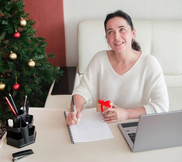 Beyaz kazaklı genç bir kadın ev ofisinde notlar yazıyor, hediye kutusunu elinde tutuyor, Noel için hazırlanıyor. Kış tatilleri indirimi.