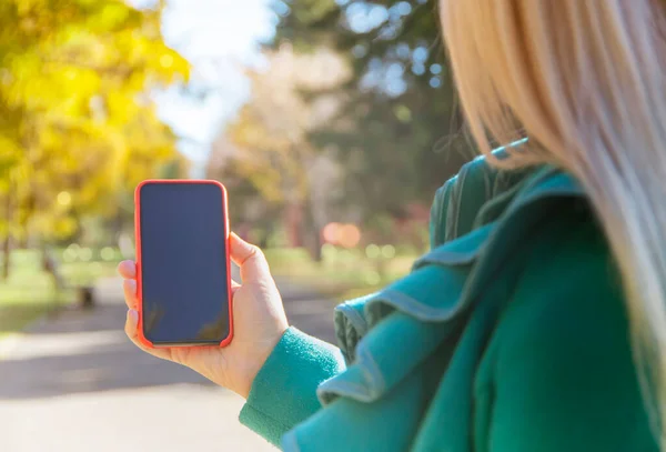 Closeup photo of female hands holding modern smartphone with blank screen. Ready layout for text message or content. Female hands with a mobile phone. Blank display.Autumn street