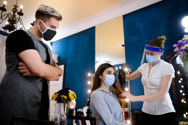 Professional hairstylists with masks on their faces, doing a hair job on a woman in a beauty salon during COVID-19 pandemic.