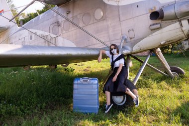 Yüzü maskeli, güzel giyimli bir kadın eski bir uçağın tekerinde bavuluyla oturuyor.