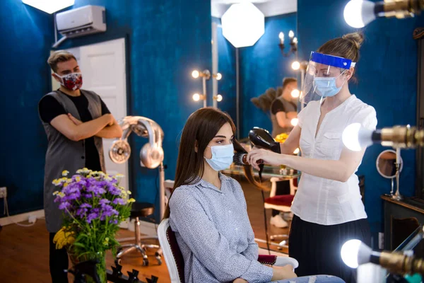 Yüzlerinde maskeler olan profesyonel kuaförler COVID-19 salgını sırasında güzellik salonunda bir kadının saçını yapıyor..