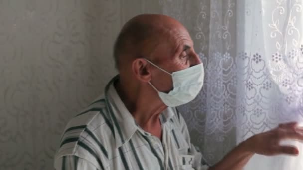 Ein älterer Rentner mit medizinischer Maske steht am Fenster eines Hauses und schaut hinaus. Ältere Menschen in Quarantäne inmitten der Coronavirus-Pandemie. Selektiver Fokus.